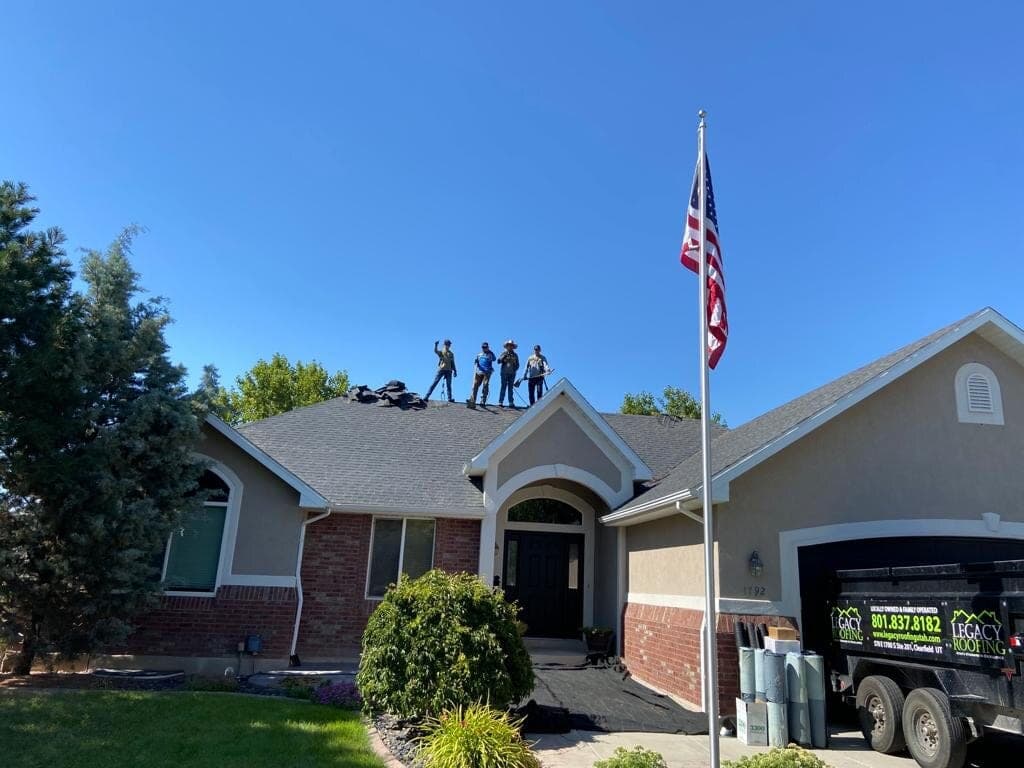 Legacy Roofing crew waving from a finished shingle roof beside a Legacy Roofing branded trailer